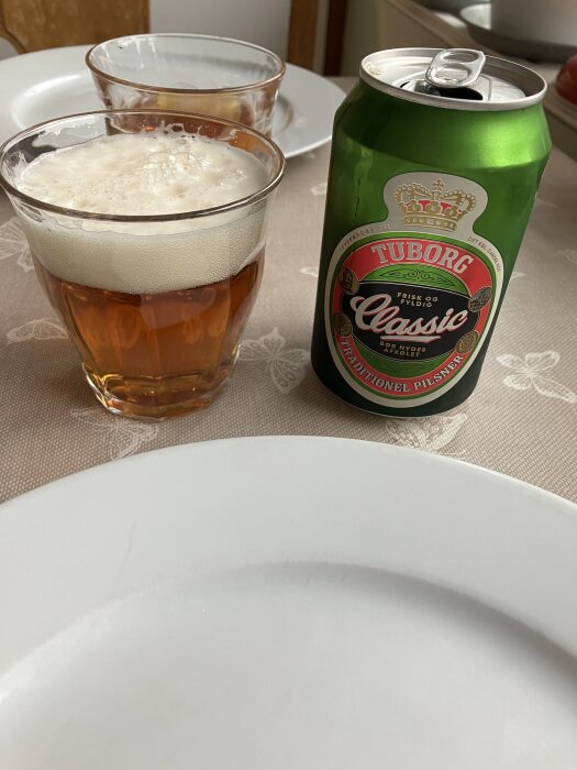 En burk Tuborg Classic öl och ett glas med skummande öl på ett dukat bord.