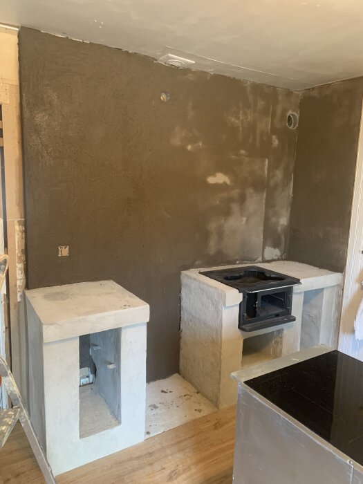 Kök under renovering med betongmurar, spishäll på plats och spiskåpa saknas. Väggar med mörk betongprimer från Byggmax.