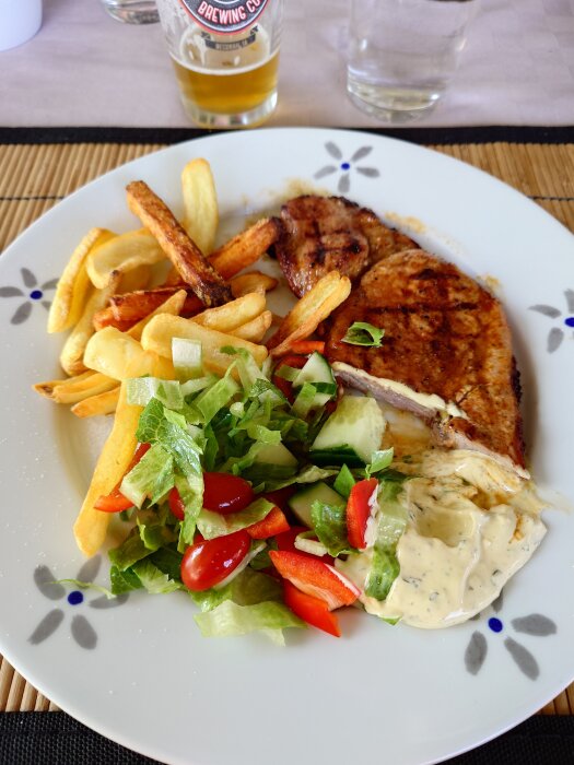 Tallrik med grillad karré, pommes frites, sallad och bearnaisesås, delvis uppätet. Glas öl i bakgrunden.