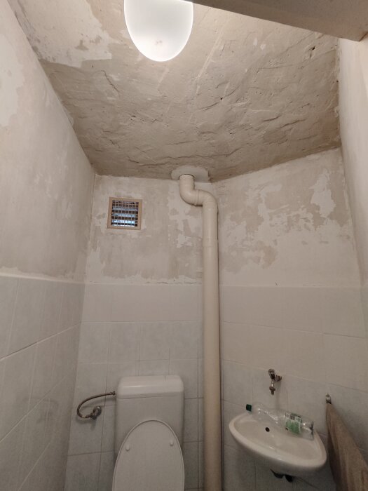 Ojämnt och flagnande tak i badrum, med synligt ventilationsrör och slitna väggar efter vattenskada. Toalett, handfat och småväggfläkt även synliga.