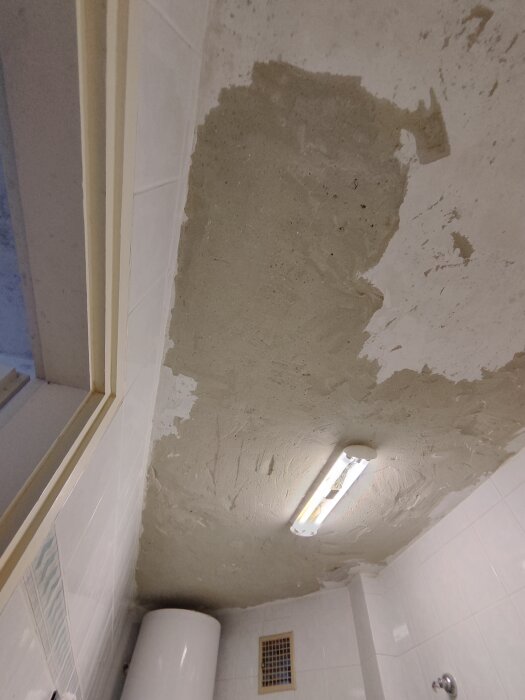 Ojämn putsad takyta i ett badrum med vita kaklade väggar, lysrör och lucka för ventilation.