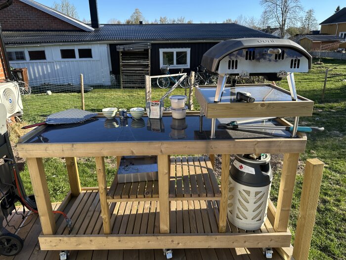 Träbänk med pizzaugn, pizzaspade, gasolflaska och bakredskap på en gräsmatta.