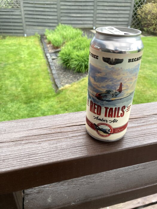 En burk Red Tails Amber Ale på ett trästaket med en grön trädgård i bakgrunden.