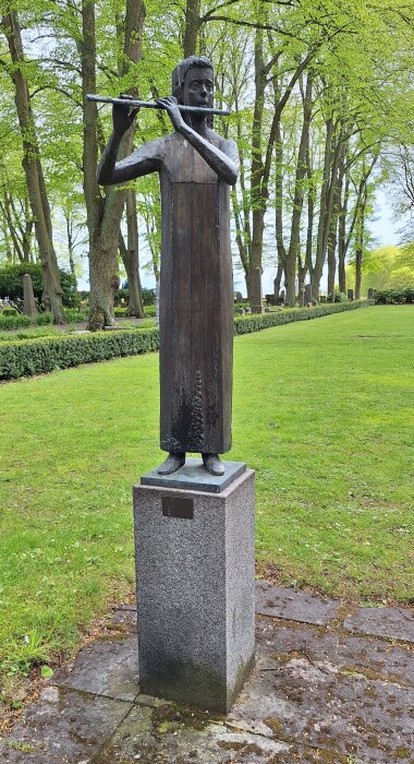 Staty av person som spelar flöjt, stående på stenfundament i grönskande park med trädallé i bakgrunden.