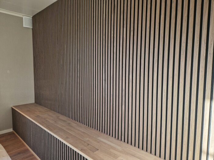 Vägg med vertikala träpaneler och en lång trähylla, redo för installation av ambient belysning och hörnuttag för ström.