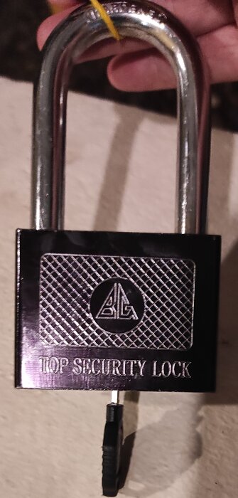 Hänglås med texten "Top Security Lock", base av metall och upphöjt emblem, håller nyckel inuti.