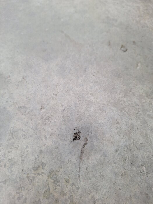 Närbild av en betongyta med små hål och ojämnheter, inklusive ett större tydligt hål i mitten, omgiven av flammiga och varierande färgade områden.