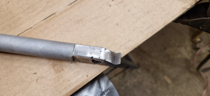 Trasigt fräshandtag i metall, reparerat med svetsning. Ligger på en arbetsbänk av trä, svårt utsatt på grund av tidigare misstag.