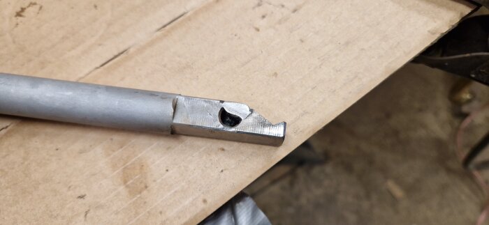 Trasigt metallhandtag från fräs, delvis avbrutet och lagat med svets, ligger på kartongskiva.