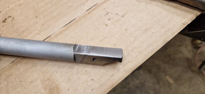 Ett metallhandtag från en fräs, svetsat och bearbetat, ligger på en kartongskiva.