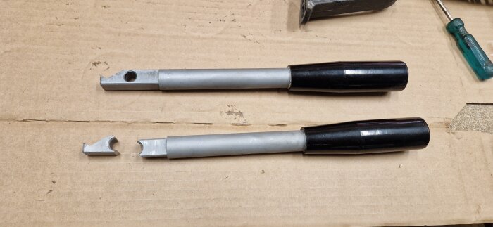 Avbrutet och helt handtag för fräs maskin ligger på en kartong, med ett avbrutet metallstycke bredvid. En skruvmejsel syns i hörnet.