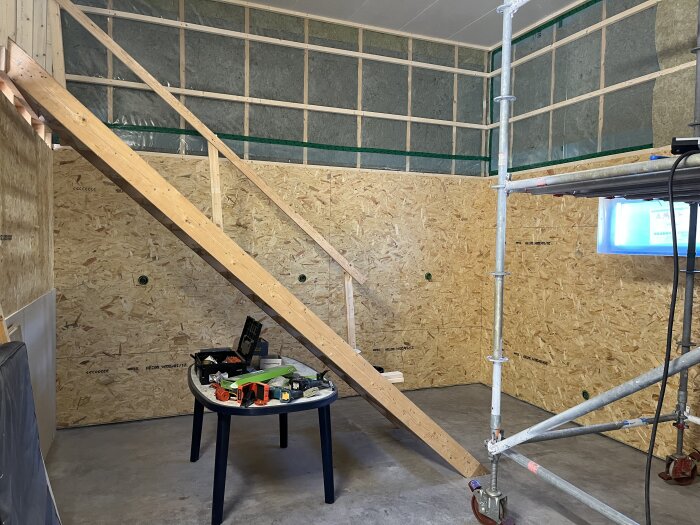 Väggar med OSB-skivor och gipsskivor, en byggställning och ett bord med verktyg i ett rum under renovering.
