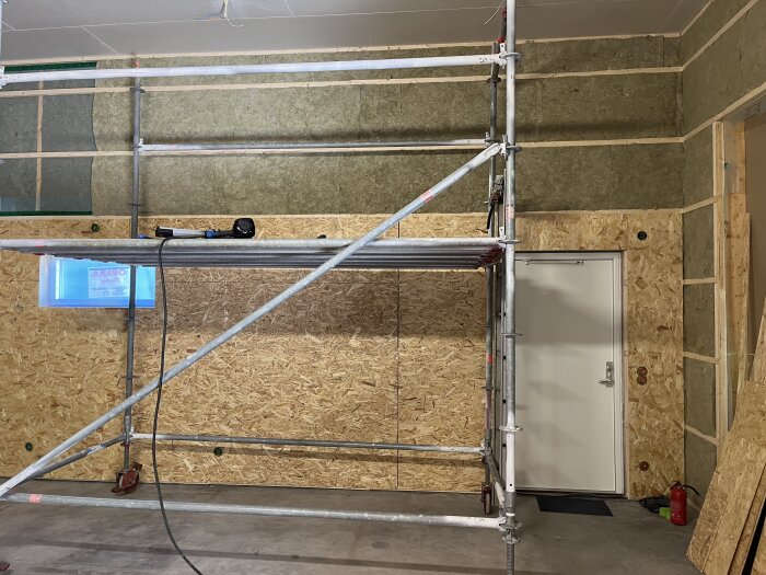 Ställning framför vägg med OSB-skivor och gips, dörr och ett fönster synliga. Verktyg och brandsläckare på golvet, under byggprojekt.