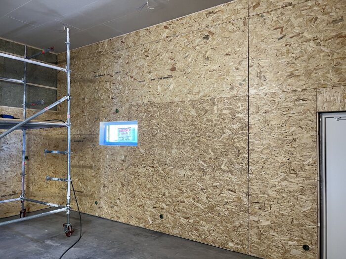 En vägg täckt med OSB-skivor och delvis gips i ett rum med byggnadsställning och ett litet fönster.