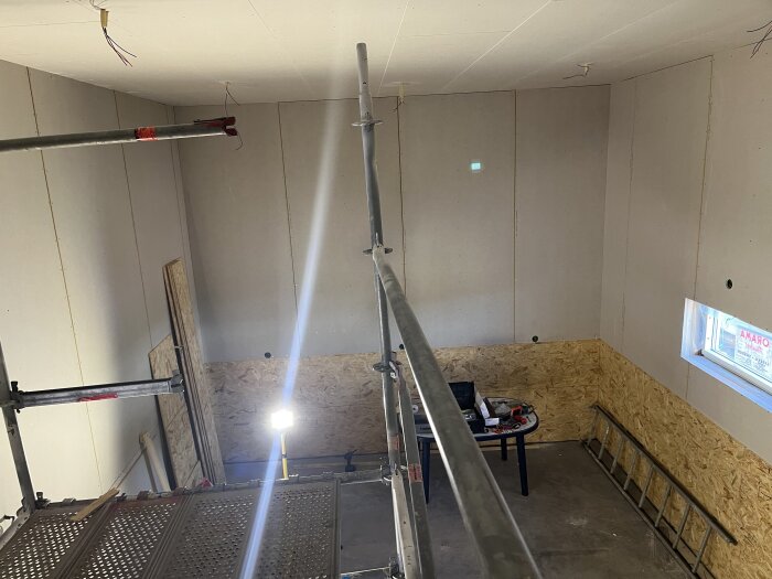 Rum under konstruktion med färdig osb och gips, liten byggställning framför och bord med verktyg i bakgrunden.