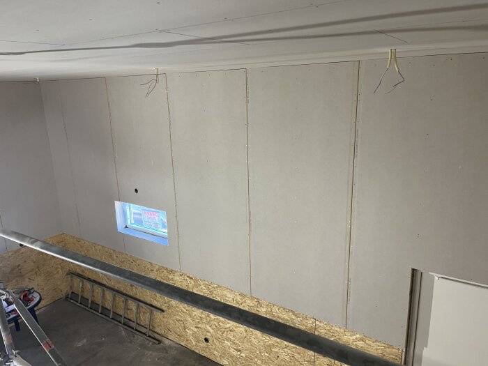 Vägg med monterat OSB och gipsskivor, liten fönsteröppning syns, stege och verktyg på golvet, elektriska kablar hänger från taket.