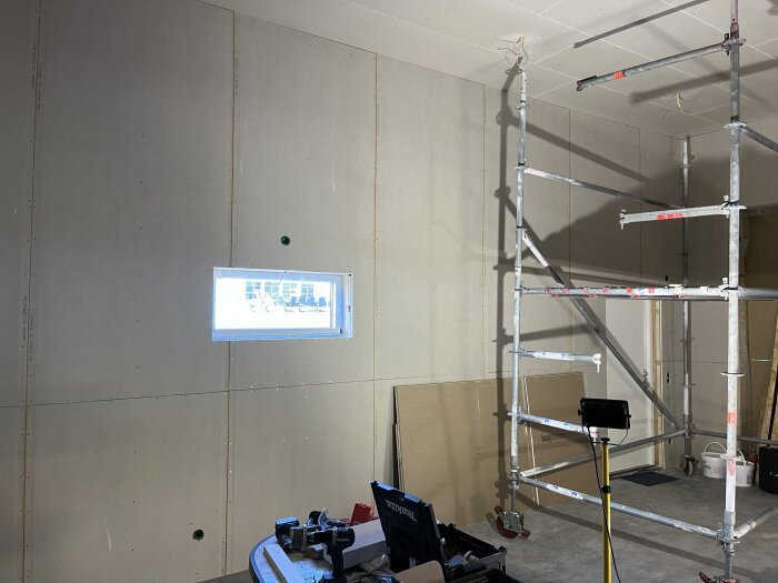 Vägg med OSB och gips färdigställd, ställning och verktyg i rummet, litet fönster på väggen.