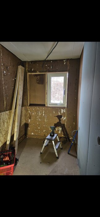 Renoveringsrum med oslipade väggar, träskivor, ett fönster, en stege med borrmaskin och en verktygslåda på golvet.