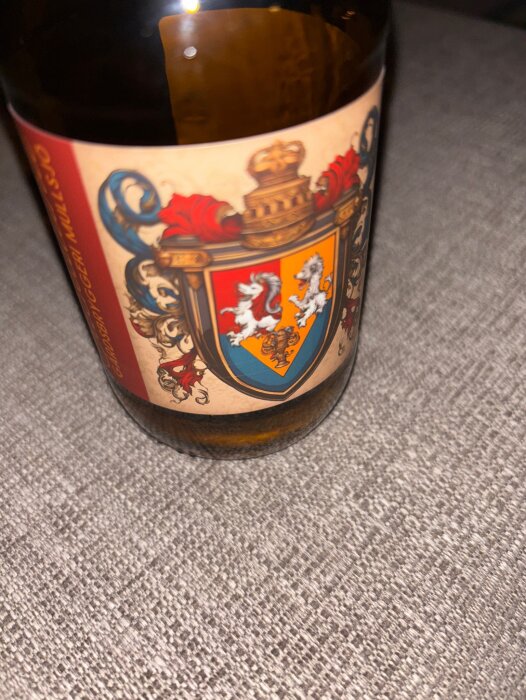 En ölflaska med etikett från Mullsjö gårdsbryggeri, visar ett färgglatt vapenmotiv och text mot en grå bakgrund.