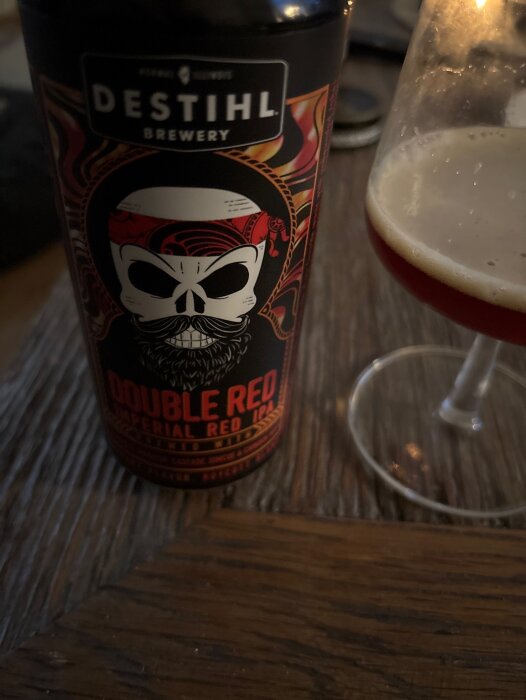 Burk med Destihl Double Red Imperial Red IPA och ett glas delvis fyllt med öl på ett träbord.