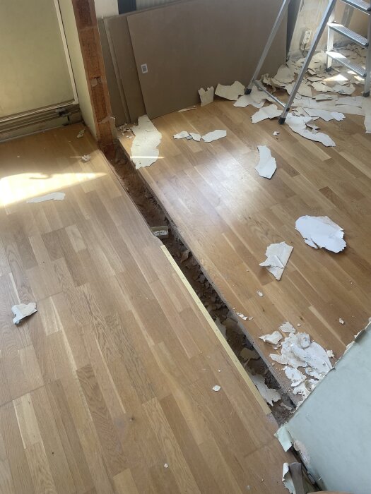 Ojämn golvnivå med råspont och parkett där en vägg har tagits bort, omgiven av skräp och en stege i ett rum.