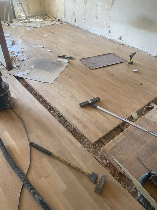 Golv med borttagen vägg visar hål och nivåskillnader, verktyg och material ligger utspridda på parkettgolv i en renoveringsmiljö.