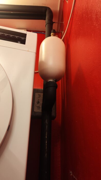Svart rör som fortsätter uppåt längs en röd vägg, bredvid en vit hushållsmaskin.