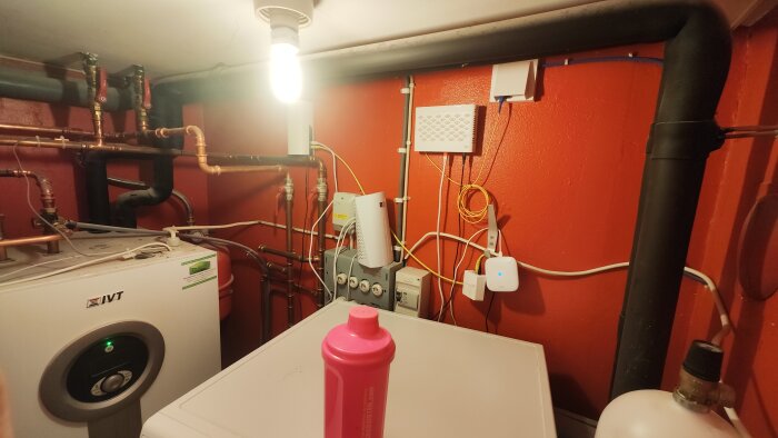 Pannrum med rörinstallationer som fortsätter uppåt, synliga kopparrör och en vit värmepump på en röd vägg.