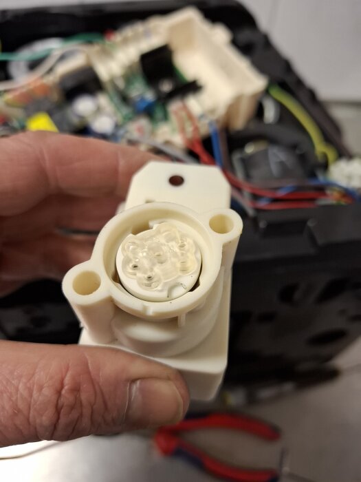 Hand håller en keramisk ventil del till en kaffemaskin för rengöring, oskärpa i bakgrunden visar elektroniska komponenter och verktyg.