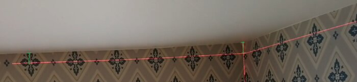 Vägg med mönstrad tapet och tak där en röd laserlinje projiceras från hörn till hörn för att mäta vinklar och lod.