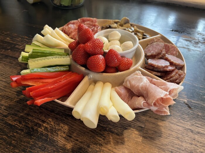 Tapasbricka med jordgubbar, gurkstavar, paprikaremsor, ost, prosciutto och salami på träbord.