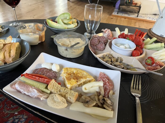 Tapas med salami, ost, parmaskinka, grönsaker, jordgubbar och melon på tallrik, med chokladask och vinglas i bakgrunden.
