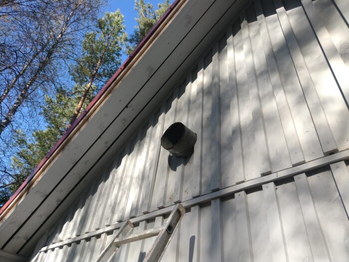 Ventilrör installerat genom yttervägg på byggnad, stege syns lutad mot väggen, omgiven av träd och blå himmel.