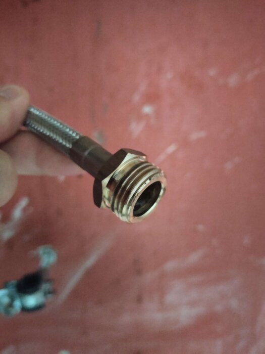 Mässingskoppling med gängor för vattenanslutning i närbild, hålls mot en röd bakgrund.