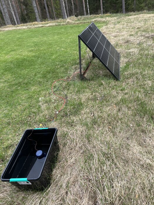 Solpanel med sladd kopplad till plastbehållare på gräsig mark nära skogskant.