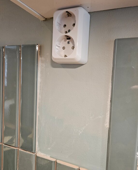 Utanpåliggande dubbelt eluttag på vägg i kök, omgiven av halvkaklade väggar med glansiga kakelplattor, under en hylla eller skåp.