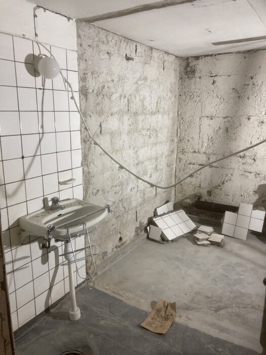 Badrum under renovering med sliten vägg, kakelplattor på golvet och ett handfat på en kaklad vägg.