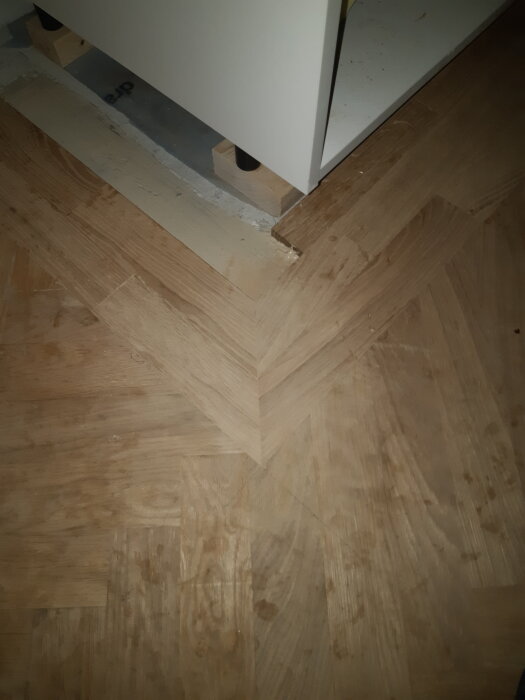 Trägolv i fiskbensmönster med en vit möbel på reglar, delvis täckt av laminat eller faner från Ikea för höjdjustering.
