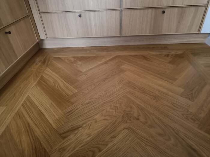 Fiskbensparkett i trä framför trälådor i ett kök, med en sockel under skåpen för att höja dem.