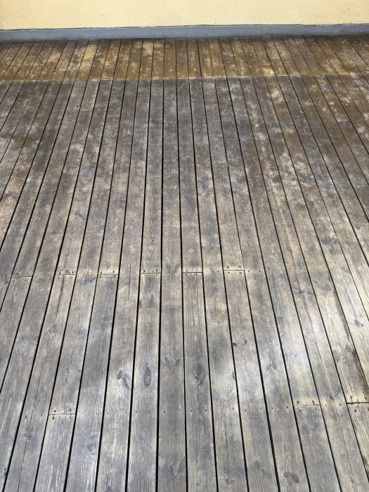 Träterrass med olika nyanser och fläckar, vissa områden mörkare. Bakgrund av ljusgul vägg och grå kant. Rengjord men fortfarande varierad färg.