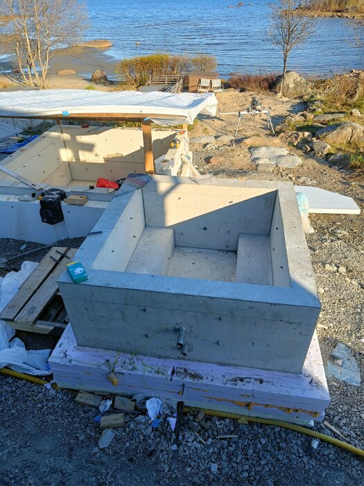 Betongbassänger under konstruktion med utsikt över en strand och hav. En täckt struktur och byggmaterial är synliga runtom.