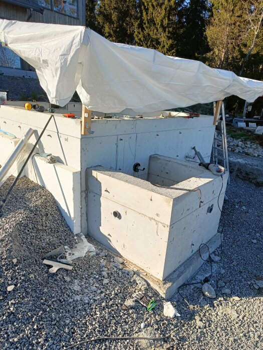 Betongstrukturer under uppbyggnad, delvis täckta av en presenning, omgivna av grus med byggverktyg och material runtomkring.