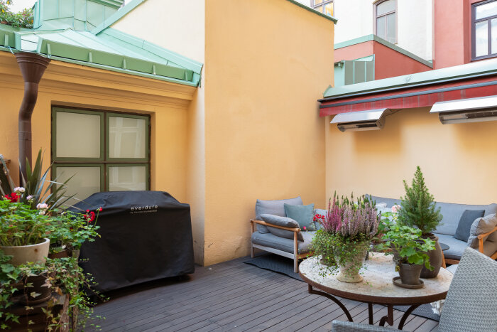 Terrass med trämöbel, grön växt, kulört växt, bord med fler krukväxter och en täckt grill mot ljusgul vägg.