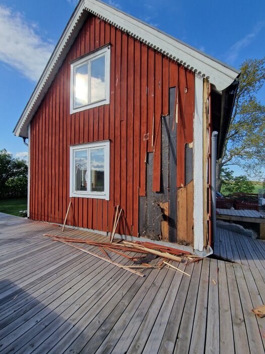 Rött hus med nyinstallerade fönster, fuktpåverkad panel delvis borttagen. Synligt trävirke och brädor på altanen, klarblå himmel i bakgrunden.