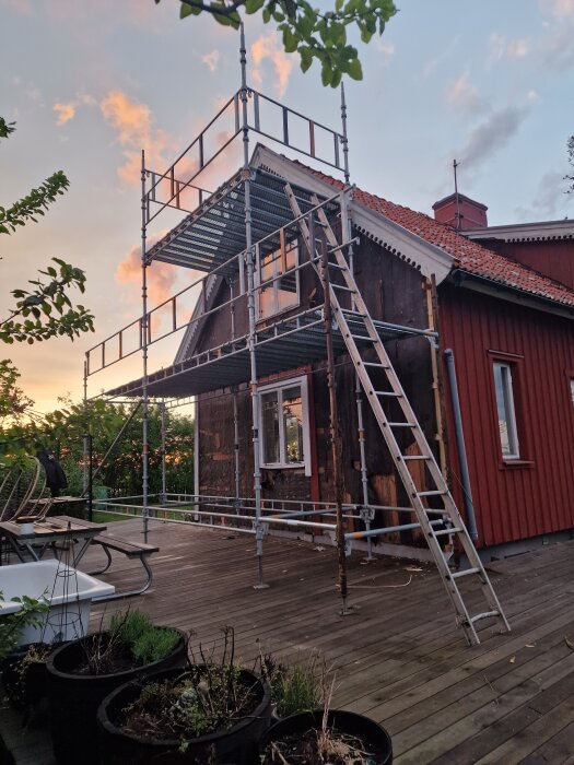 Byggställning uppställd vid ett hus med röda träväggar och nytt fönster. Gamla träpaneler på gaveln borttagna, läkt synlig. Stege lutar mot ställningen.
