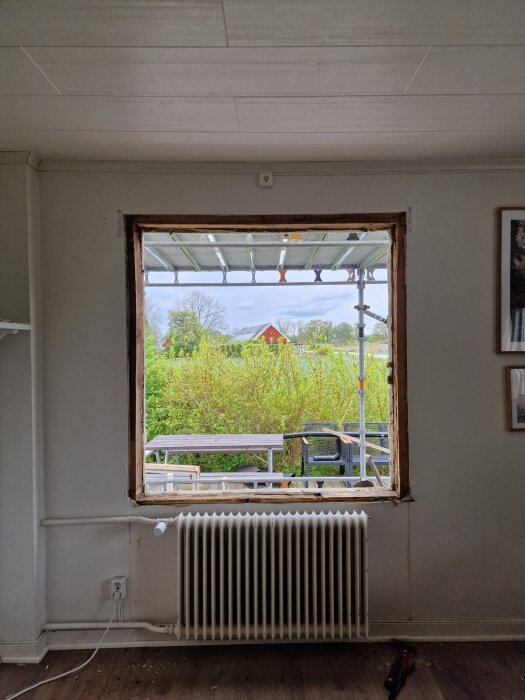 Ett hål i en vägg där ett fönster ska monteras, med en utsikt över en trädgård och byggnadsställning utanför. En element under hålet.