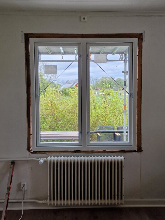 Nya fönster installerade i en vitmålad vägg, med en utsikt över en grön trädgård och byggnadsställning utanför.