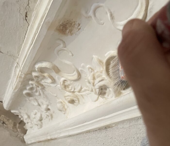 En hand målar en gipsstukatur på en vägg med en pensel; detaljerade blommönster och skador syns på stukaturen.