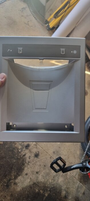 Lock med monteringsskruvhål och ikoner för ljus och glob, hålls i handen, ligger på verkstadsgolv med cykelpedal i bakgrunden.