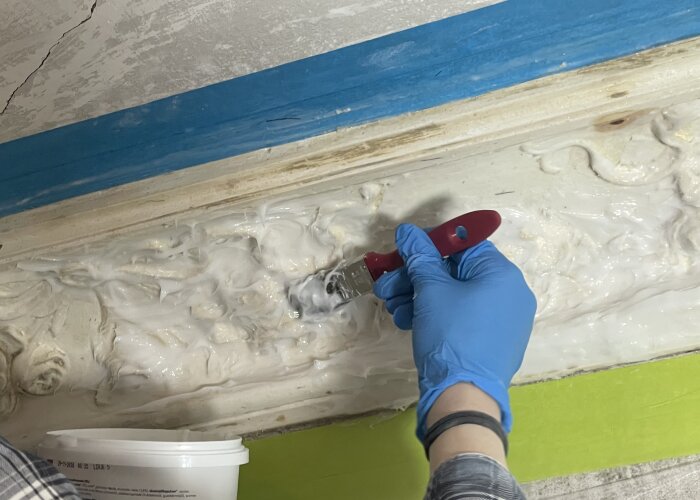 Person applicerar silikon med pensel på gipsstukatur, iklädd blå handske; del av renoveringsprojekt.